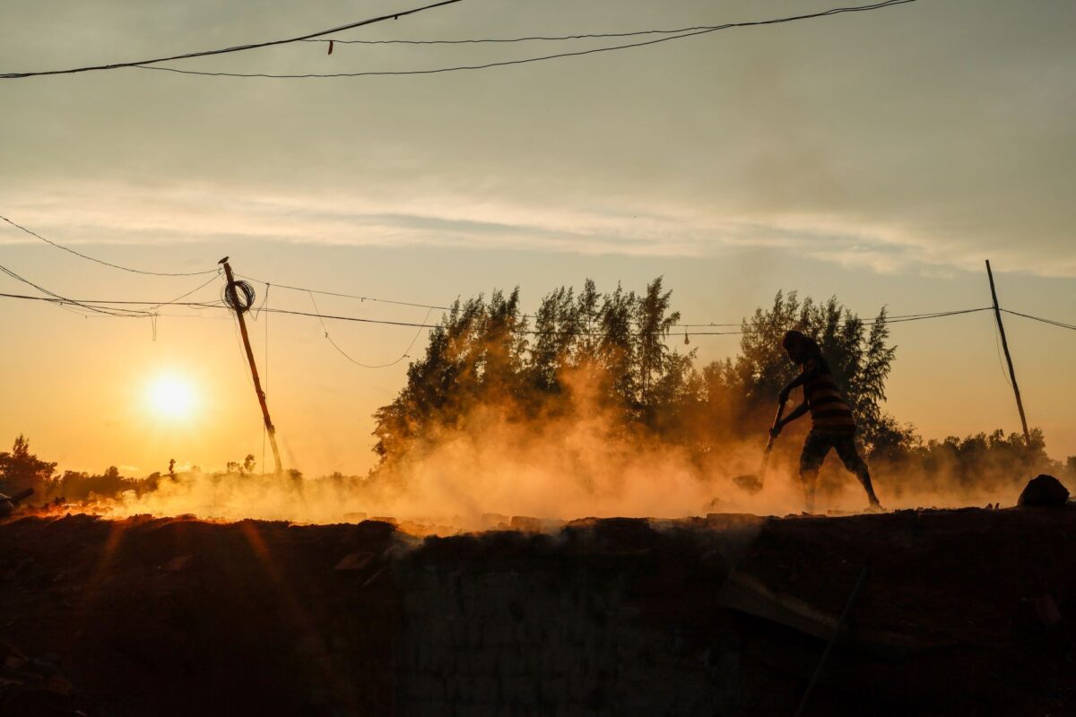 A worker digging amidst rising dust at sunset with power lines and trees contrasting against the sky.